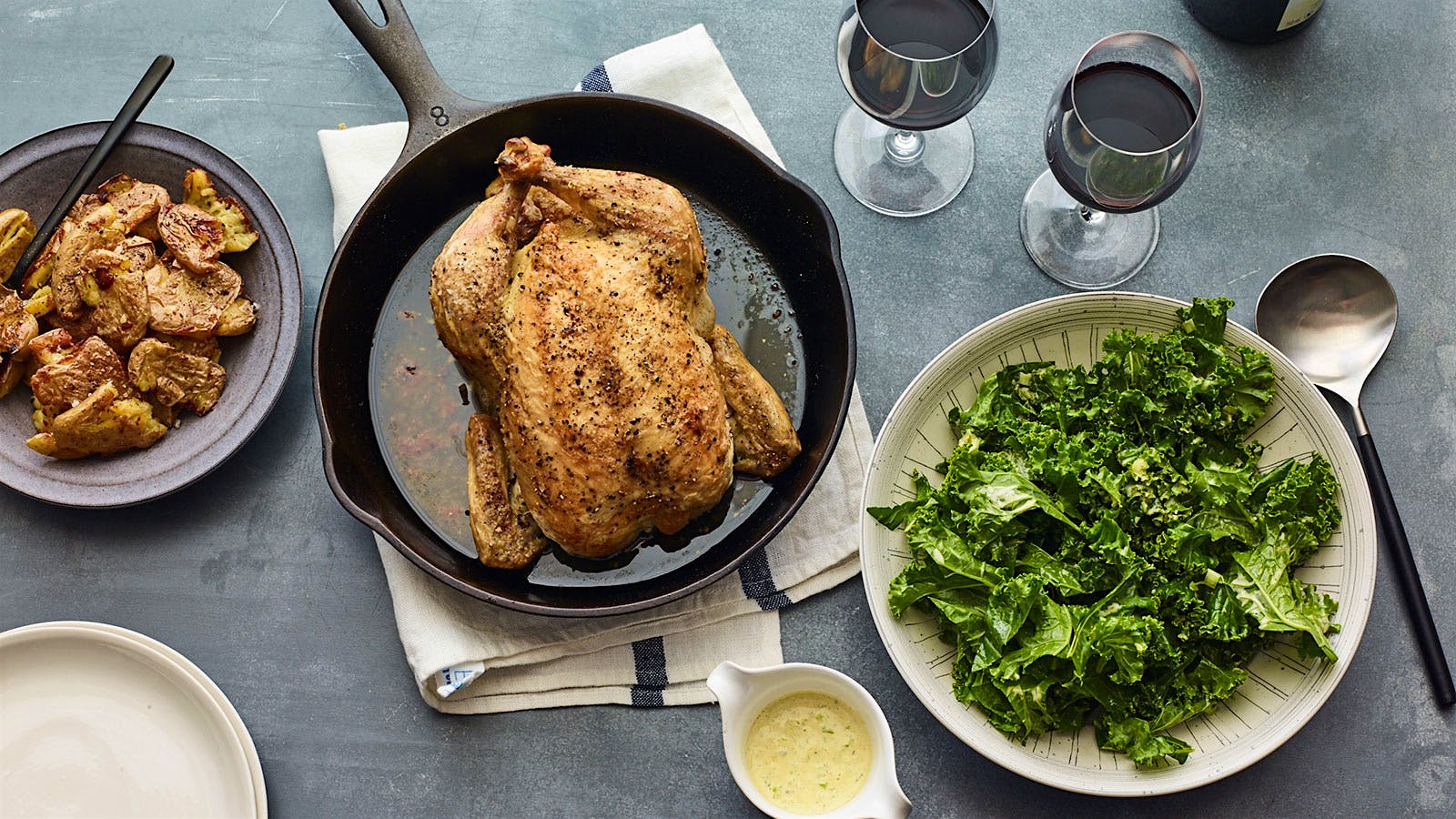 A roast chicken in a cast iron pan accompanied by smashed roast potatoes, salad greens with a cup of dressing and two glasses of red wine