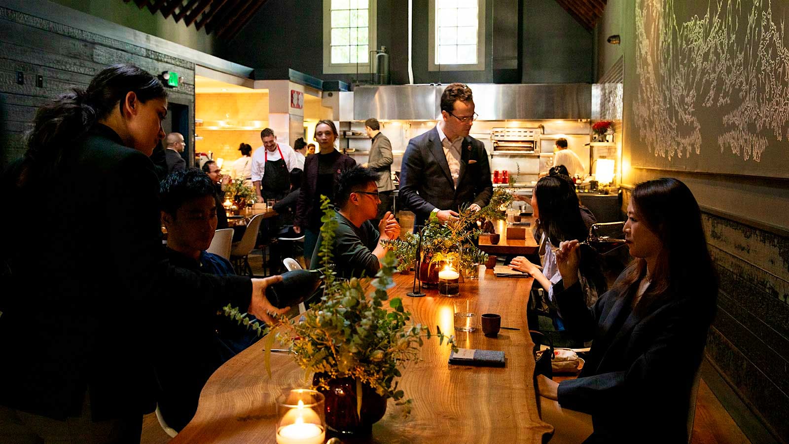 The dining room at Lazy Bear with guests seated around a long communal wood table and the open kitchen in the background