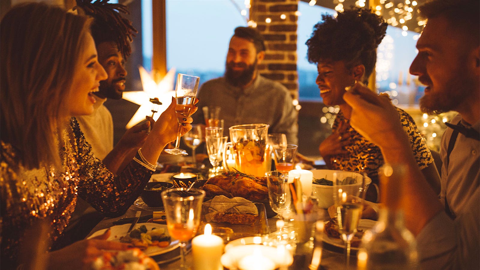 A group of friends laughing around a dinner table, with glowing lights hanging around them