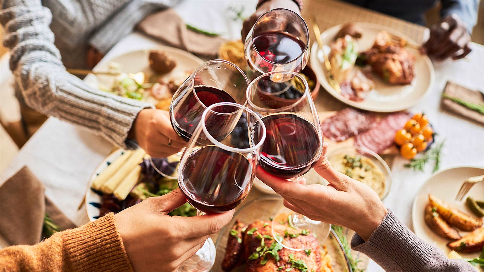 People clinking glasses of red wine over a table set for a Thanksgiving meal, with turkey and other plates